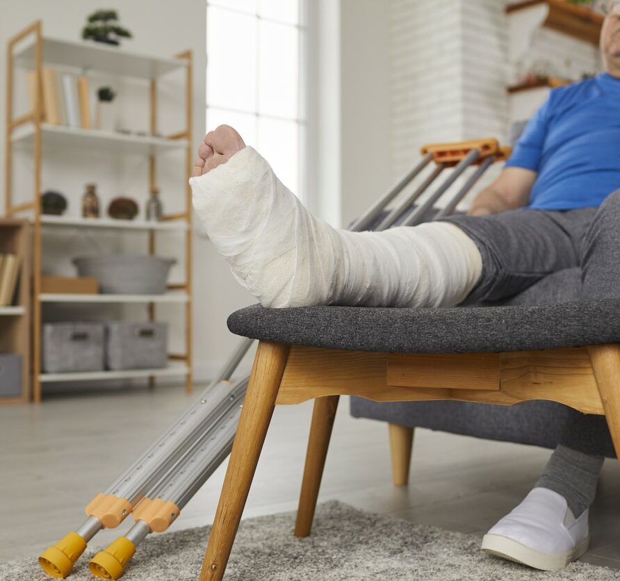 Senior man with broken leg in plaster cast sitting on couch and talking on phone