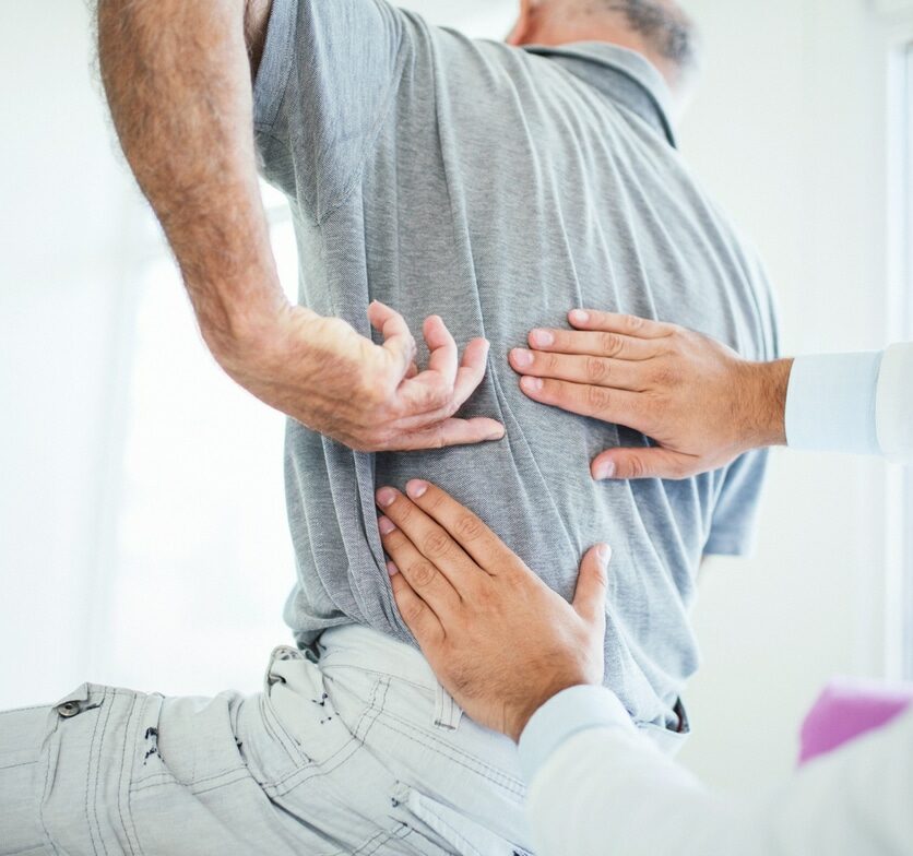 Closeup rear low angle view of an early 60's senior gentleman having some back pain. He's at doctor's office having medical examination by a male doctor. The patient is pointing to his lumbar region.